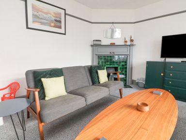 A living room with a gray sofa green cushions a wooden coffee table a fireplace with green tiles a dresser and a television at Chilton in Bude