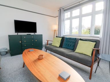 A living room with a gray sofa with green and blue cushions a wooden coffee table a dark green cabinet with a television and a floor lamp at Chilton in Bude