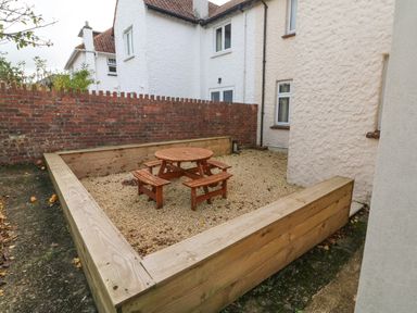 An outdoor patio area with a round wooden picnic table and benches on gravel surrounded by wooden edging at Chilton in Bude