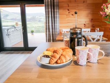 A kitchen table with croissants jam and butter a French press and cups in a wooden cabin with a view to a deck and hills at Bryn Eiddon Log Cabin in Machynlleth