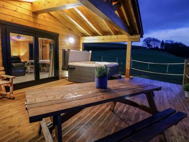 A wooden outdoor patio with a picnic table a hot tub and glass doors leading inside at Bryn Eiddon Log Cabin in Machynlleth