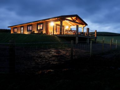 A wooden cabin with exterior lights on and a porch in a grassy area at Bryn Eiddon Log Cabin in Machynlleth