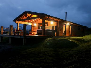 An illuminated wooden cabin with a covered porch and outdoor seating at Bryn Eiddon Log Cabin in Machynlleth