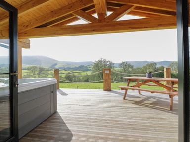 A wooden deck with a hot tub and picnic table overlooking green fields and hills at Bryn Eiddon Log Cabin in Machynlleth