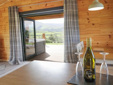 A wooden interior room with a wine bottle and two glasses on a table next to an open door showing a deck and green hills at Bryn Eiddon Log Cabin in Machynlleth