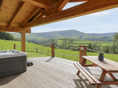 A wooden deck with a hot tub and picnic table overlooking green hills at Bryn Eiddon Log Cabin in Machynlleth