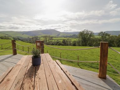 A wooden picnic table with a potted plant on a deck overlooking green fields and distant hills at Bryn Eiddon Log Cabin in Machynlleth