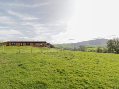 A wooden cabin on a grassy hill with a fence and distant hills in the background at Bryn Eiddon Log Cabin in Machynlleth