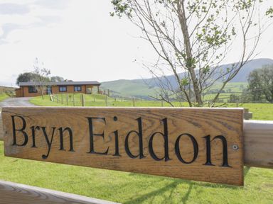 A wooden sign with the text Bryn Eiddon in front of a fenced grassy area with a wooden cabin and hills in the background at Bryn Eiddon Log Cabin in Machynlleth