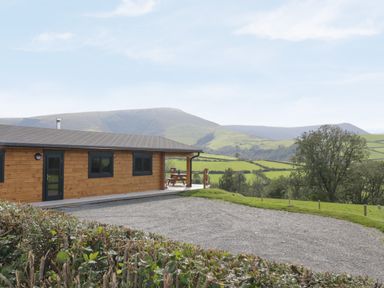 A wooden log cabin with a gravel driveway and a picnic bench on a porch with hills and trees in the background at Bryn Eiddon Log Cabin in Machynlleth