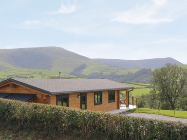 A wooden cabin with a gravel path and hedge in front with hills in the background at Bryn Eiddon Log Cabin in Machynlleth