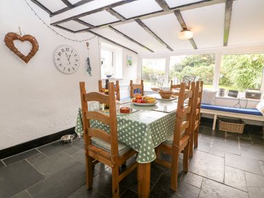 A dining room with a wooden table covered with a green polka dot tablecloth and wooden chairs next to large windows with garden view at Tyddyn Serri in Rhosneigr