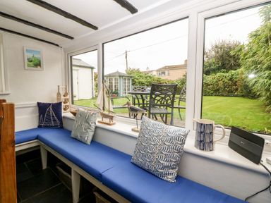 A small seating area with blue cushions and nautical pillows by large windows overlooking a garden with outdoor table and chairs at Tyddyn Serri in Rhosneigr