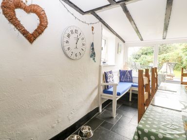 A dining area with a corner bench with blue cushions and pillows a clock on the wall wooden chairs around a table with polka dot tablecloth and windows showing a garden at Tyddyn Serri in Rhosneigr