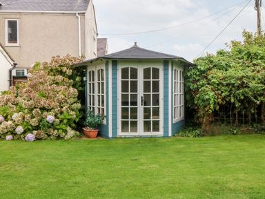 A small blue garden house with white framed windows and doors surrounded by plants and grass at Tyddyn Serri in Rhosneigr