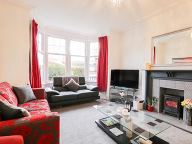 A living room with a red patterned sofa and a black sofa near a bay window with red curtains and a fireplace with a television at Beach House in Bridlington