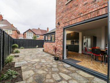 A paved backyard with plants along a black fence and an open door to a kitchen with a dining table and chairs at Beach House in Bridlington