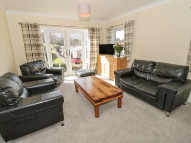 A living room with black leather sofas a wooden coffee table a wooden cabinet with a television flowers and checked curtains at Arwelfa in Penmaenmawr