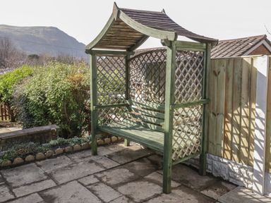 A wooden garden bench with a lattice back and sides on a paved patio next to a wooden fence at Arwelfa in Penmaenmawr