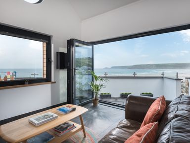 A sitting area with a leather sofa and orange cushions next to a wooden coffee table and a balcony overlooking the beach at The Old Beach Store in Sennen Cove