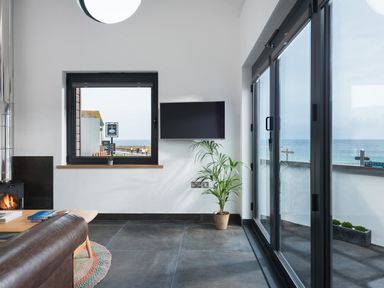 A living room with a fireplace a wall mounted TV a plant a window and glass doors showing a sea view at The Old Beach Store in Sennen Cove