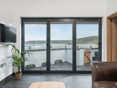 A living room with a television a plant and a leather sofa facing glass doors with a view of a beach at The Old Beach Store in Sennen Cove