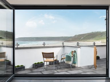 A balcony with two chairs and a blanket overlooking a beach and hills at The Old Beach Store in Sennen Cove
