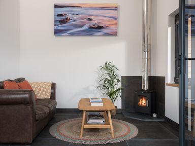 A living room with a brown leather sofa a wooden coffee table with books a round woven rug a potted plant and a wood burning stove at The Old Beach Store in Sennen Cove
