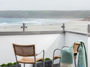 A balcony with two woven chairs and a green blanket overlooking a beach at The Old Beach Store in Sennen Cove