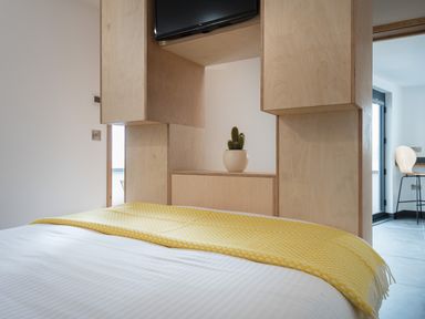 A bedroom with a yellow blanket on the bed a wooden partition with a potted cactus and a television in the room at The Old Beach Store in Sennen Cove