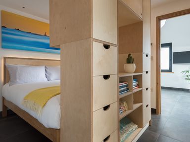 A bedroom with a wooden bed and white bedding next to a wooden shelving unit with drawers and books at The Old Beach Store in Sennen Cove