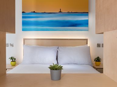 A bedroom with a bed with white pillows and sheets a plant on a wooden surface and a large painting of a seascape at The Old Beach Store in Sennen Cove