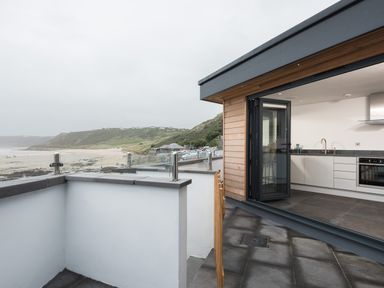 A patio with glass and metal railing overlooking a beach and hills next to a kitchen with white cabinets at The Old Beach Store in Sennen Cove