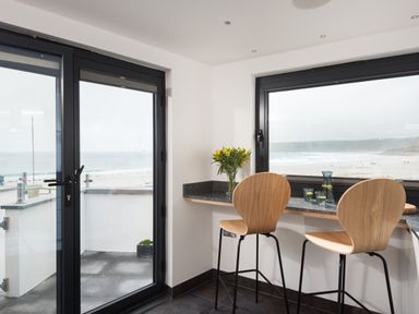 A kitchen corner with two wooden bar stools flowers and glassware on the countertop by large windows overlooking the beach at The Old Beach Store in Sennen Cove