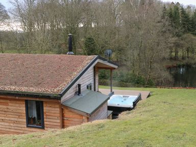 A wooden cabin with a pebble roof and a hot tub outside near trees and a body of water at Tarn Lodge in Bowness-On-Windermere