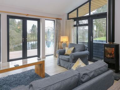 A living room with grey sofas a wooden coffee table a lamp and a fireplace with glass doors overlooking trees and a river at Tarn Lodge in Bowness-On-Windermere