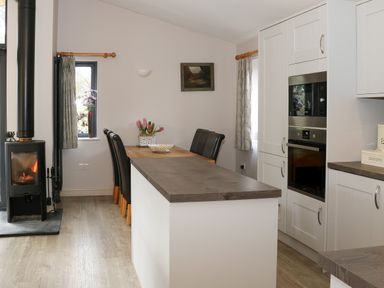 A kitchen area with a wood-burning stove a wooden table with chairs and white cabinets at Tarn Lodge in Bowness-On-Windermere