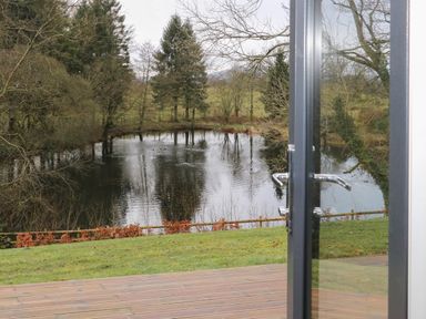 A view through glass doors showing a wooden deck grass area a pond and trees at Tarn Lodge in Bowness-On-Windermere