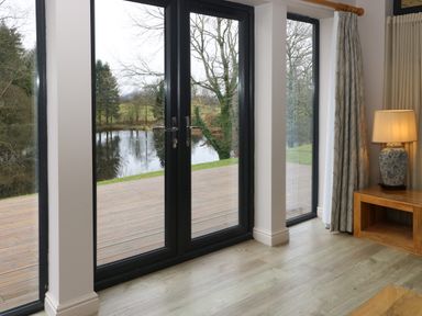 Large black-framed glass doors and windows opening to a wooden deck with a view of trees and water with a wooden side table with a ceramic lamp and curtains at Tarn Lodge in Bowness-On-Windermere