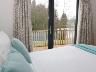 A bedroom with a bed and blue pillows facing large glass doors opening to a balcony overlooking trees and water at Tarn Lodge in Bowness-On-Windermere