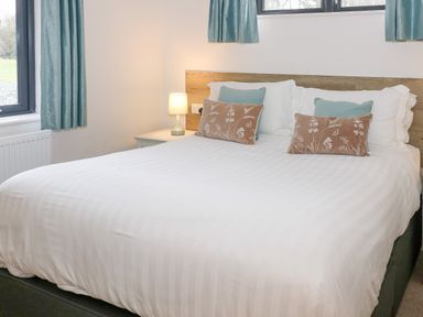 A bedroom with a white bed, teal and beige pillows, a wooden headboard, a bedside table with a lamp, and teal curtains at Tarn Lodge in Bowness-On-Windermere