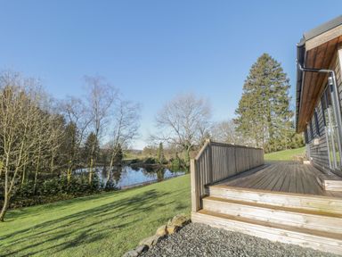 A wooden deck with steps overlooking a grassy area with trees and a pond at Tarn Lodge in Bowness-On-Windermere