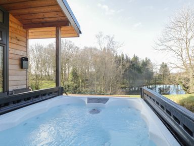 An outdoor hot tub on a wooden deck overlooking trees and a small pond at Tarn Lodge in Bowness-On-Windermere