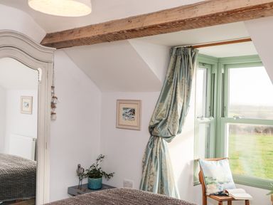 A bedroom corner with a window and green curtains a wooden chair with a cushion and an open book a bedside table with a plant and a bird figurine and a wall mirror at Ty ar Ben Bryn in Pencarnisiog near Rhosneigr