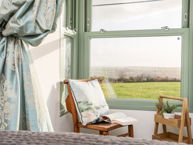 A wooden chair with a cushion and an open book near a window with green frames and curtains and a plant on a wooden stand at Ty ar Ben Bryn in Pencarnisiog near Rhosneigr