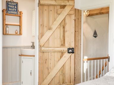 A wooden door partially open showing part of a bathroom with a sink and a wooden shelf and a hallway with a staircase and a small sailboat decoration on the wall at Ty ar Ben Bryn in Pencarnisiog near Rhosneigr