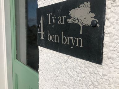 A house number plaque with the text 4 Ty ar ben bryn and a tree symbol on a textured white wall next to a green door at Ty ar Ben Bryn in Pencarnisiog near Rhosneigr