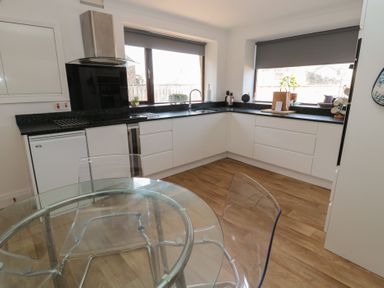 A kitchen with white cabinets black countertops a glass dining table and two clear chairs at Changing Tide in Beadnell