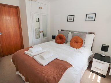 A bedroom with a bed dressed with white and orange linens a bedside table with a lamp a wardrobe with mirrored doors and a wooden door at Changing Tide Beadnell