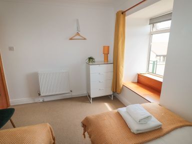 A bedroom with two single beds with orange covers a white chest of drawers with a lamp and plant a window seat with a wooden surface and yellow curtains at Changing Tide in Beadnell